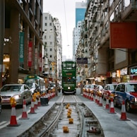 A busy urban street with a classic green tram in the center, surrounded by high-rise buildings. The street includes colorful shops and signs on either side. Red and white traffic cones line the tram tracks, and various vehicles, including taxis, are parked along the side.