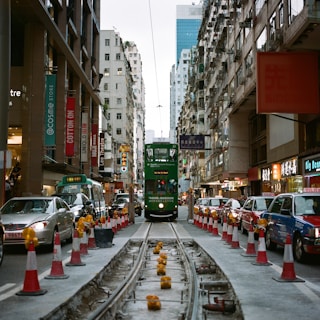 A busy urban street with a classic green tram in the center, surrounded by high-rise buildings. The street includes colorful shops and signs on either side. Red and white traffic cones line the tram tracks, and various vehicles, including taxis, are parked along the side.