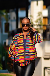 A person stands confidently outdoors wearing a vibrant, colorful striped jacket paired with black sunglasses and earrings. Their pose exudes confidence and style, with one hand placed in a pocket. The background is softly blurred, suggesting a modern urban environment with hints of greenery.