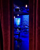 A dimly lit room with a stage-like setting, featuring a row of round tables and chairs. Blue lighting casts a cool ambiance on the brick wall backdrop, with a soft glow from scattered light fixtures. Heavy curtains partially conceal the scene, evoking a sense of mystery and anticipation.