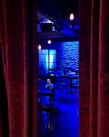A dimly lit room with a stage-like setting, featuring a row of round tables and chairs. Blue lighting casts a cool ambiance on the brick wall backdrop, with a soft glow from scattered light fixtures. Heavy curtains partially conceal the scene, evoking a sense of mystery and anticipation.