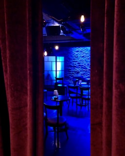 A dimly lit room with a stage-like setting, featuring a row of round tables and chairs. Blue lighting casts a cool ambiance on the brick wall backdrop, with a soft glow from scattered light fixtures. Heavy curtains partially conceal the scene, evoking a sense of mystery and anticipation.