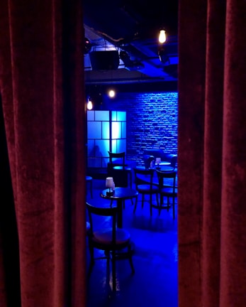 A dimly lit room with a stage-like setting, featuring a row of round tables and chairs. Blue lighting casts a cool ambiance on the brick wall backdrop, with a soft glow from scattered light fixtures. Heavy curtains partially conceal the scene, evoking a sense of mystery and anticipation.