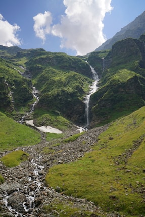 A lush, green mountain landscape with a cascading waterfall flowing down the rocky terrain. The sky is clear with some fluffy white clouds drifting above.