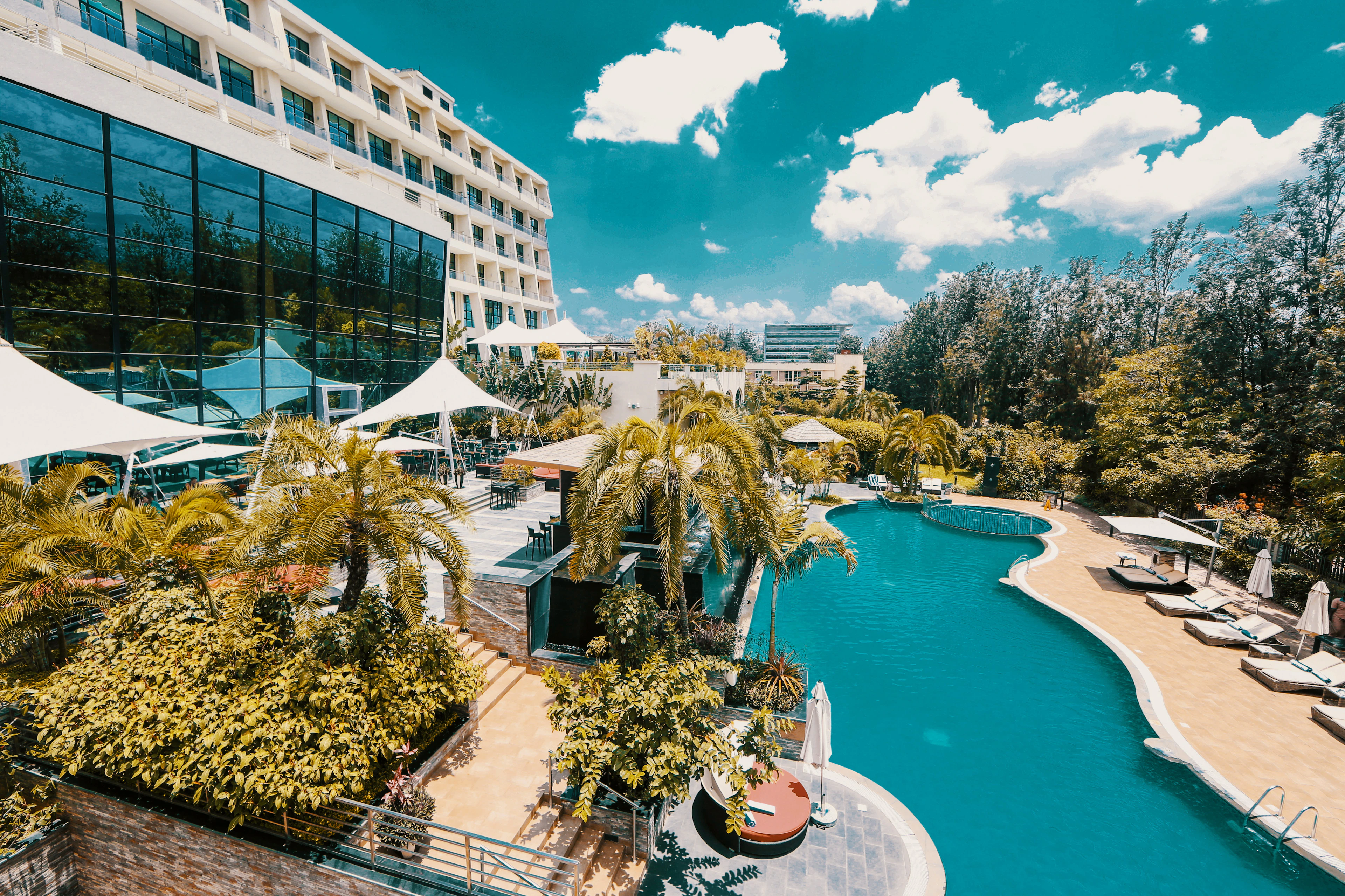 The curved white facade and palm-fringed swimming pool terrace of the Marriott hotel in Kigali, with sun loungers and parasols beside the water