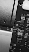 A monochrome image featuring a digital watch with a rectangular face, displaying the time and date against a black design blueprint. Next to it is a closed black notebook with a subtle embossed logo and a white AirPods case. Part of a laptop with an aluminum finish is visible at the bottom left corner.