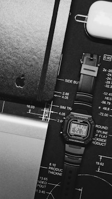 A monochrome image featuring a digital watch with a rectangular face, displaying the time and date against a black design blueprint. Next to it is a closed black notebook with a subtle embossed logo and a white AirPods case. Part of a laptop with an aluminum finish is visible at the bottom left corner.