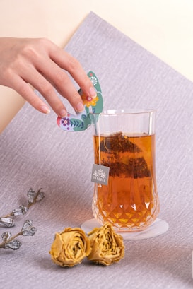 A hand delicately holds a colorful, floral-themed butterfly card above a faceted glass filled with amber-colored tea. Two dried yellow roses lie on a textured gray surface next to the glass. The tag attached to the glass reads 'TEACORA'.