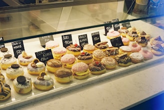 A display case filled with a variety of doughnuts, each topped with different icings, sprinkles, and toppings such as chocolate pieces and cookies. The doughnuts are arranged neatly, each with a small price tag placed in front of them.