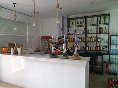 A modern bar setup with a counter displaying decorative hookahs. Behind the counter, shelves are stocked with various bottles of alcoholic beverages and some glassware. There's a red popcorn machine to the side. The setting includes potted plants, a fruit basket, and hanging rope lights above.