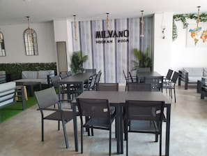 A modern hookah lounge interior with sleek black tables and chairs arranged neatly. The walls have a vertical striped pattern, and there's decorative foliage, including potted plants and wall vines. A sign reading 'Milvano Hookah Room' is mounted on the wall. The atmosphere is clean and minimalist, with some contemporary lighting fixtures hanging from the ceiling.