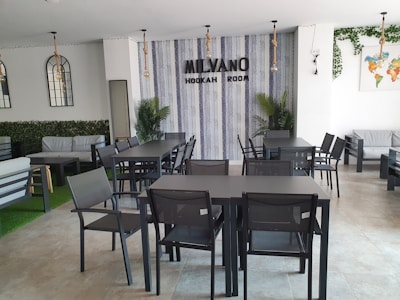 A modern hookah lounge interior with sleek black tables and chairs arranged neatly. The walls have a vertical striped pattern, and there's decorative foliage, including potted plants and wall vines. A sign reading 'Milvano Hookah Room' is mounted on the wall. The atmosphere is clean and minimalist, with some contemporary lighting fixtures hanging from the ceiling.