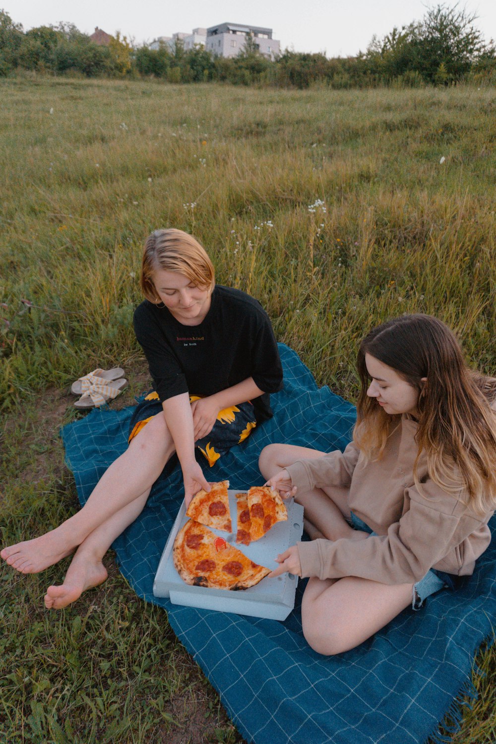 two girls sitting on a blanket eating pizza