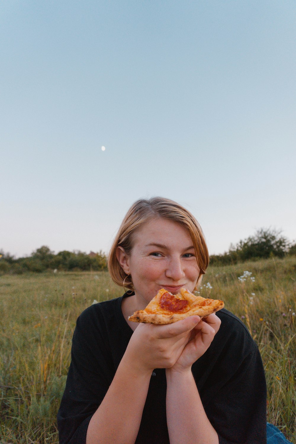 a woman sitting in a field eating a slice of pizza