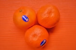 Three round, orange fruits are placed on a textured orange surface. Each fruit has a blue sticker labeled 'sweetC'. The natural and vivid color of the fruits stands out against the backdrop.