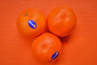 Three round, orange fruits are placed on a textured orange surface. Each fruit has a blue sticker labeled 'sweetC'. The natural and vivid color of the fruits stands out against the backdrop.