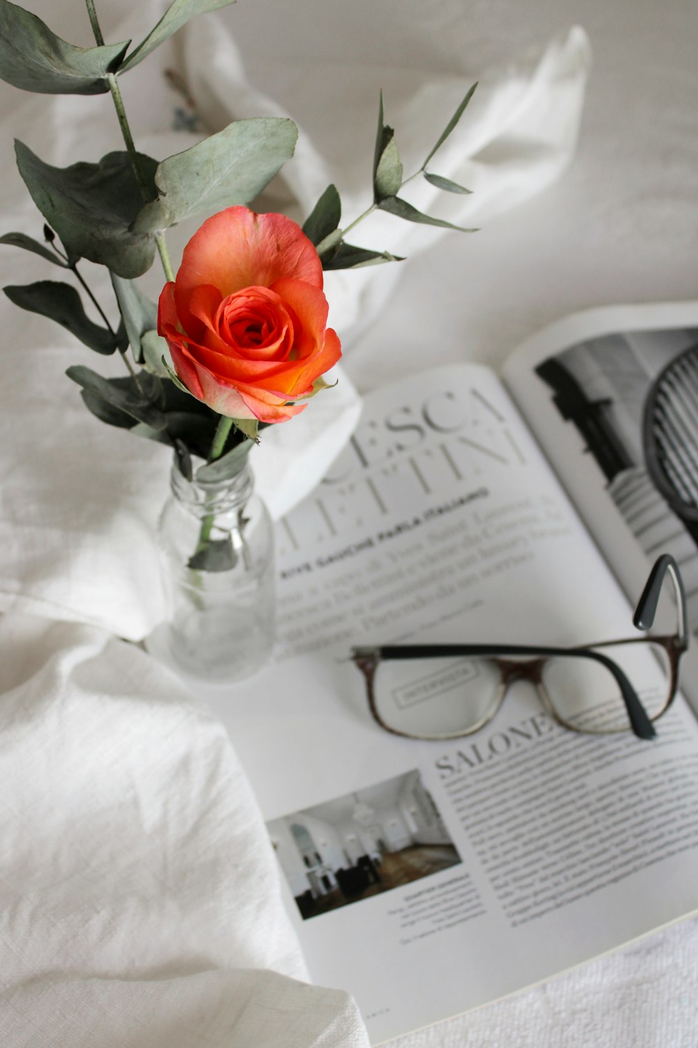 eine Vase mit einer Rose und Brille auf einem Bett