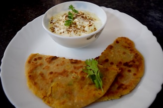 A plate with two pieces of flatbread accompanied by a bowl of yogurt garnished with herbs. The flatbread appears to be spiced and cooked to a golden brown, and is placed on a white plate. The yogurt is sprinkled with a spice, likely cumin or pepper, and decorated with fresh cilantro leaves.