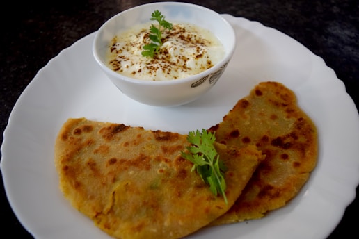 A plate with two pieces of flatbread accompanied by a bowl of yogurt garnished with herbs. The flatbread appears to be spiced and cooked to a golden brown, and is placed on a white plate. The yogurt is sprinkled with a spice, likely cumin or pepper, and decorated with fresh cilantro leaves.
