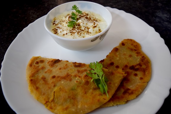 A single piece of Indian-style flatbread on a white plate, accompanied by a small bowl of creamy yogurt dip and a fresh side salad, all set on a rustic wooden table with natural daylight and soft shadows.