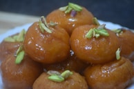 A plate of round, golden-brown sweets garnished with sliced pistachios. The sweets have a shiny, syrupy glaze, suggesting they are a type of dessert.