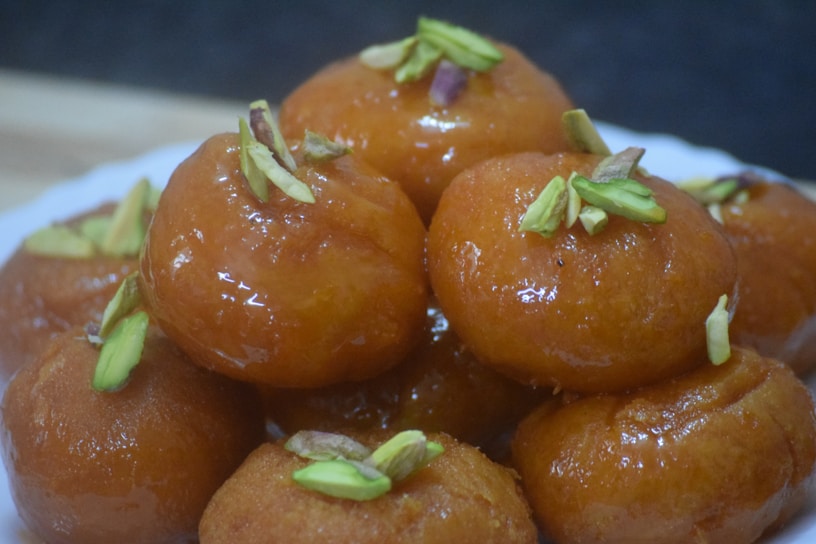 A plate of round, golden-brown sweets garnished with sliced pistachios. The sweets have a shiny, syrupy glaze, suggesting they are a type of dessert.