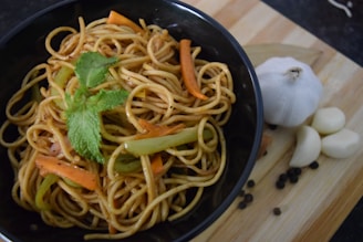 A dark bowl contains a serving of noodles mixed with slices of carrots and bell peppers, topped with a sprig of fresh mint. Beside the bowl, there are garlic cloves and black peppercorns scattered on a wooden surface, along with a whole garlic bulb.