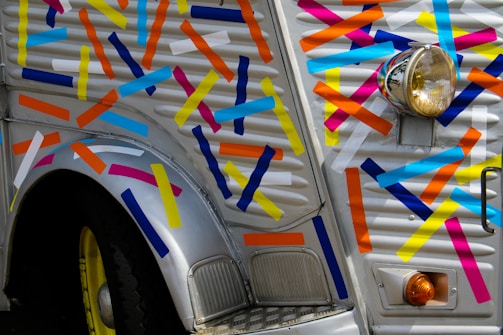A vintage vehicle covered in colorful, diagonally placed strips of tape. The body is primarily silver with a yellow wheel and a prominently affixed circular headlight. The vibrant colors include blue, yellow, orange, pink, and white, creating a playful, artistic look.
