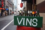 A city street scene featuring a green crate labeled 'VINS' holding several empty wine bottles. The crate rests on a red barrel, situated on the sidewalk. In the background, there are several pedestrians walking along the street, with shops and high-rise buildings lining the road. Red signage hangs from one of the buildings, adding a vibrant touch to the urban atmosphere.