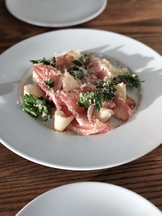 A plate of food featuring thinly sliced cured meat, possibly prosciutto, arranged with shavings of Parmesan cheese and garnished with fresh green herbs. The dish is presented on a white round plate with a wooden table surface providing a warm contrast.