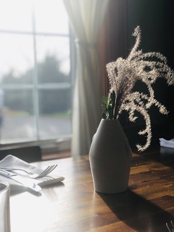 A sunlit wooden table with a white vase containing dry, wispy grass and small green leaves. Soft, diffused light streams through a large window with sheer curtains, casting gentle shadows. A neatly folded white napkin lies next to the vase, accompanied by a fork and knife.