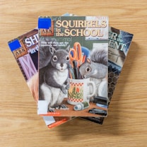 Three children's books are stacked on a wooden surface. The book on top features an illustration of two squirrels surrounded by school supplies, including scissors and a pencil, with the title 'Squirrels in the School'.