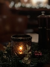 A warm, candlelit setting with a decorative candle holder featuring intricate patterns. Surrounding the candle are festive elements, including artificial leaves with a glittery finish and hints of greenery. In the dim background, blurred lights create a soft bokeh effect, adding a cozy ambiance.