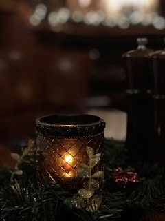 A warm, candlelit setting with a decorative candle holder featuring intricate patterns. Surrounding the candle are festive elements, including artificial leaves with a glittery finish and hints of greenery. In the dim background, blurred lights create a soft bokeh effect, adding a cozy ambiance.