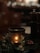 A warm, candlelit setting with a decorative candle holder featuring intricate patterns. Surrounding the candle are festive elements, including artificial leaves with a glittery finish and hints of greenery. In the dim background, blurred lights create a soft bokeh effect, adding a cozy ambiance.