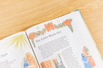 An open children's book is laid out on a wooden surface, featuring a story titled 'The Little Match Girl.' The page is adorned with colorful illustrations, including a decorative wreath with orange bows at the top. On the left, there's a drawing resembling a nativity scene and radiant star. On the right, there are small illustrations of people in winter attire standing near a brick wall.