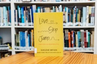 A yellow book with the title 'Love Skip Jump' is displayed on a wooden table in front of a blurred background of shelves filled with various books.