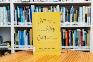 A yellow book with the title 'Love Skip Jump' is displayed on a wooden table in front of a blurred background of shelves filled with various books.