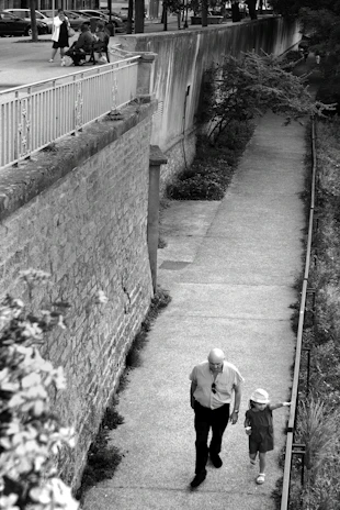 grayscale photo of man in black jacket and pants walking on pathway
