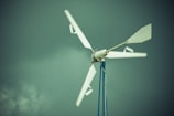 A wind turbine with white blades is mounted on a tall blue pole against a backdrop of a cloudy, greenish sky.