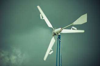 A wind turbine with white blades is mounted on a tall blue pole against a backdrop of a cloudy, greenish sky.