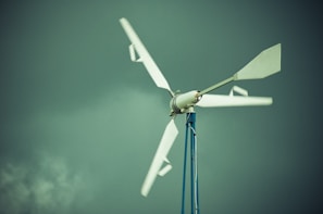 A wind turbine with white blades is mounted on a tall blue pole against a backdrop of a cloudy, greenish sky.