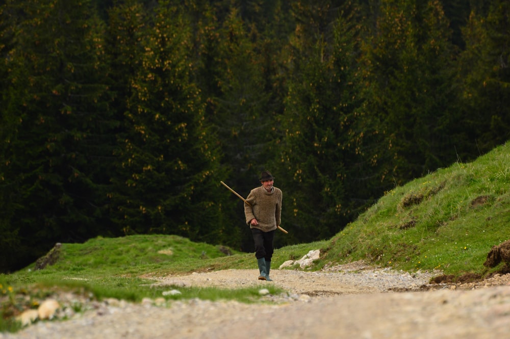 a man walking down a dirt road holding a stick