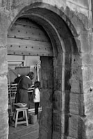 A black and white photograph depicting a woman and a child inside a stone structure with a large wooden door. The woman appears to be demonstrating or working on a loom while the child observes. There is a small stool and a basket nearby.