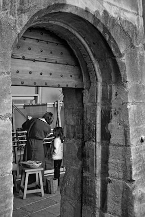 A black and white photograph depicting a woman and a child inside a stone structure with a large wooden door. The woman appears to be demonstrating or working on a loom while the child observes. There is a small stool and a basket nearby.