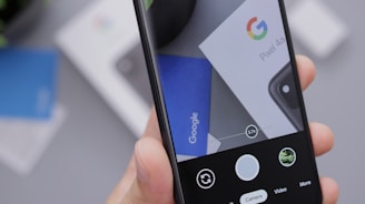 A person holding a smartphone displaying its camera interface, with an image of Google branding and Pixel 4a packaging visible on the phone screen. The scene includes a hand partially in view, focusing on a close-up of the phone. Background elements are blurred, suggesting a desk or flat surface environment.