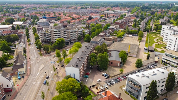 An aerial view of a suburban area featuring a mix of modern and traditional architecture. The streets are lined with trees, and a variety of buildings are visible, including residential homes, apartment complexes, and commercial structures. Vehicles are parked in designated areas, and roads are primarily empty, suggesting a calm day.