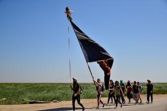 A group of people is walking along a road holding a large black flag. The scene takes place in a rural landscape with a clear blue sky and green fields in the background. The group appears to be on a journey, with some members wearing hats and carrying items.