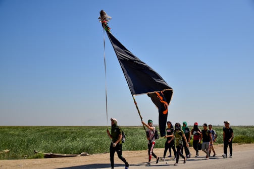 A group of people is walking along a road holding a large black flag. The scene takes place in a rural landscape with a clear blue sky and green fields in the background. The group appears to be on a journey, with some members wearing hats and carrying items.