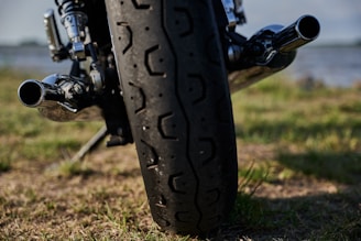 A close-up view of a motorcycle's rear tire, focusing on the detailed tread pattern. The chrome exhaust pipes are visible, reflecting light. The background consists of a blurred natural scenery with grass and a body of water, suggesting an outdoor setting.